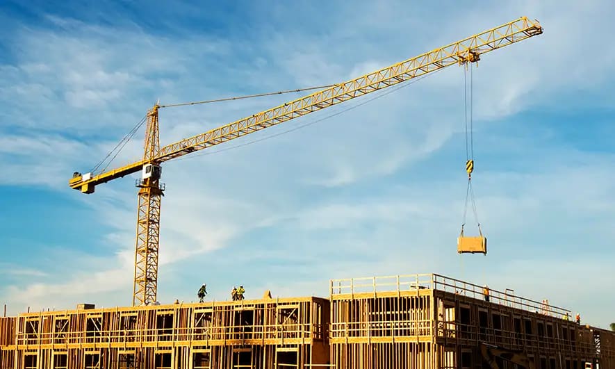 Yellow crane lifting a wooden pallet at a construction site with a partially built structure under a clear blue sky.