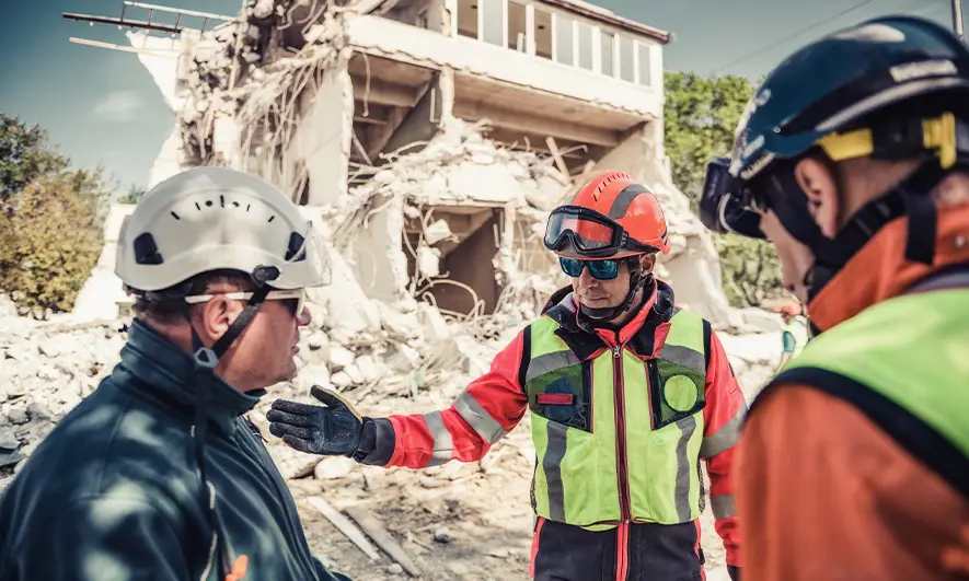 Three rescue workers in helmets and safety gear standing in front of a partially collapsed building, discussing the situation.