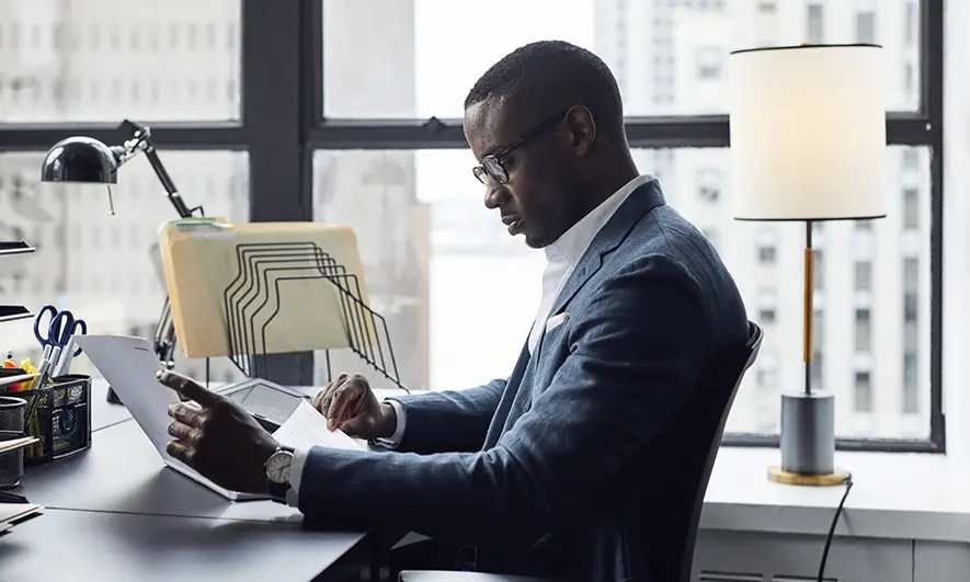 Professional seated at a desk reviewing documents in an office with large windows.