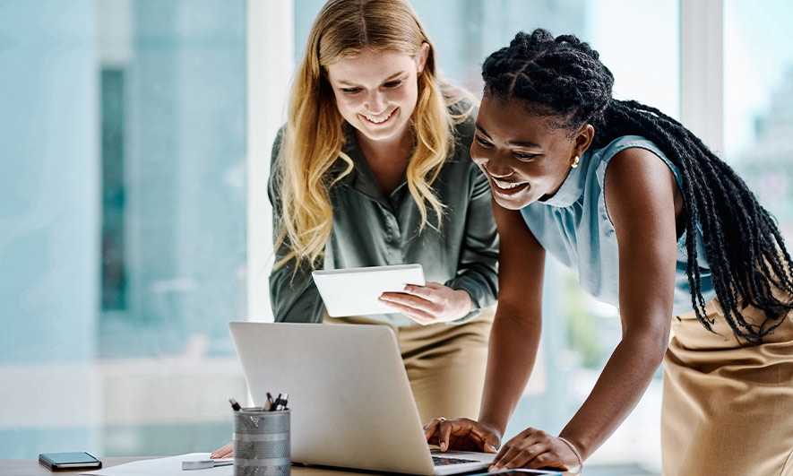 Two diverse businesswomen working together on a tablet and laptop.