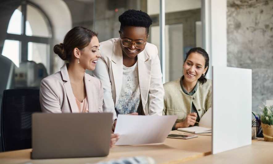 Three smiling women discuss small business mentoring while reviewing documents at a desk.