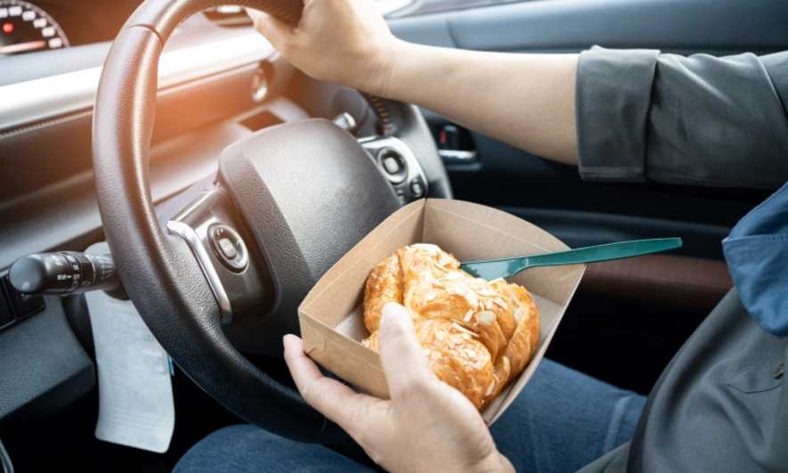 A driver holding a pastry in one hand and the steering wheel in the other