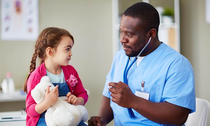 A nurse with a stethoscope sits next to a child patient holding a teddy bear.