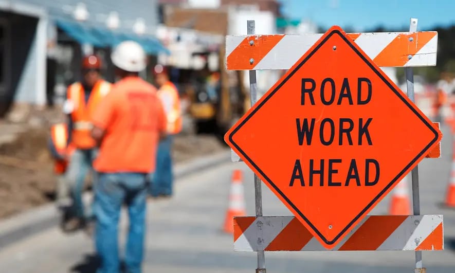 Orange ‘Road Work Ahead’ sign in the foreground with a marked construction zone and workers visible in the background.