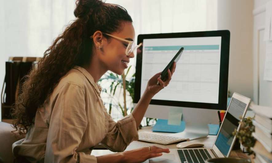 Woman working on laptop and phone at desk, exploring home business ideas with spreadsheets on screen.