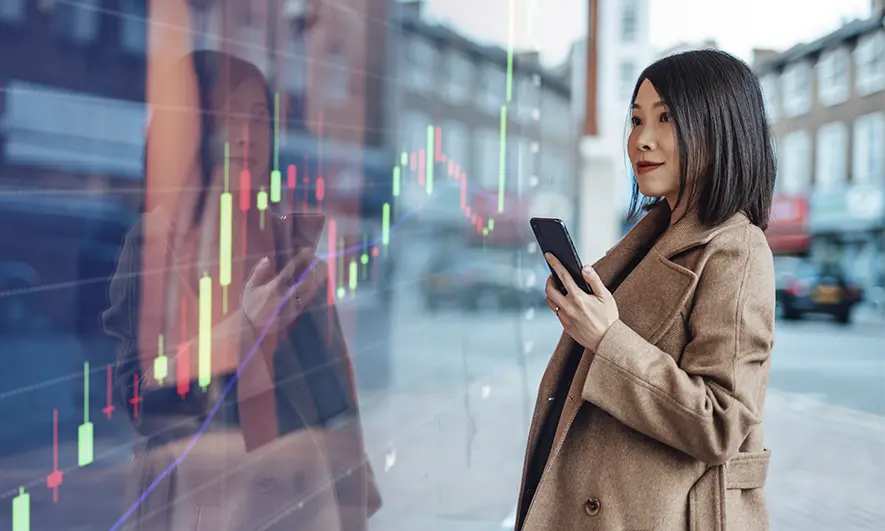 Person using a smartphone on a city street, seen through glass overlaid with a financial market chart