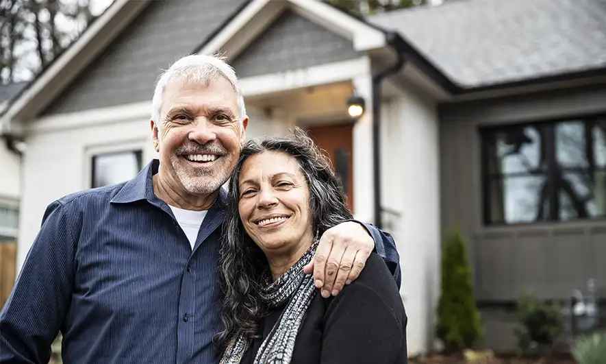 A middle-aged couple smiles in front of their home